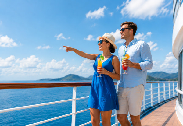 A person in a blue sleeveless dress and a light-colored hat stands beside another person wearing a light blue button-up shirt and shorts on the deck of a cruise ship. Both are holding colorful drinks and wearing sunglasses. The person in the dress is pointing toward the horizon over a bright blue ocean, with distant green hills visible under a partly cloudy sky. The ship’s white railing and deck flooring run along the right side of the scene.